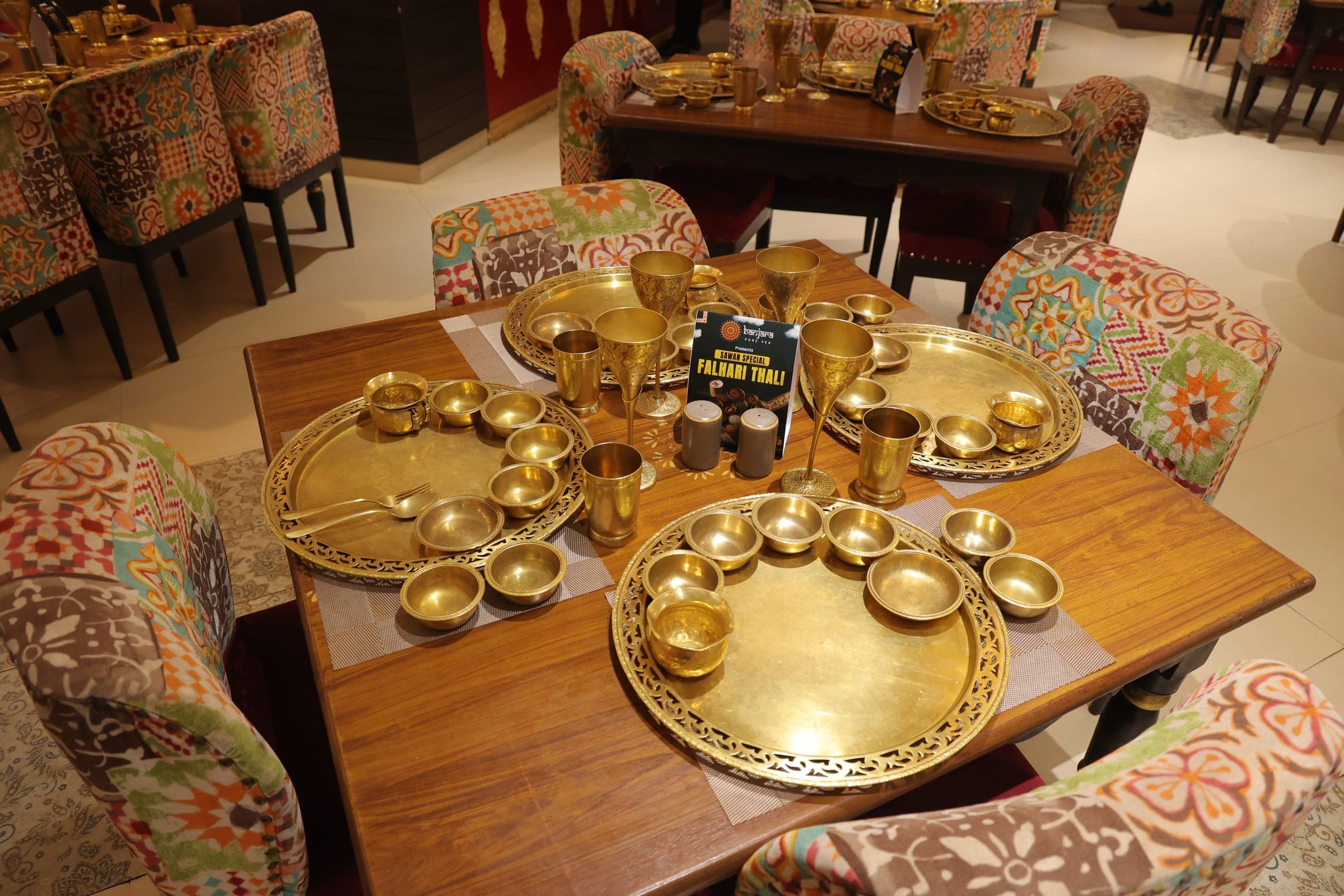 Brass thalis, goblets and colourful Banjara chairs around a wooden dining table